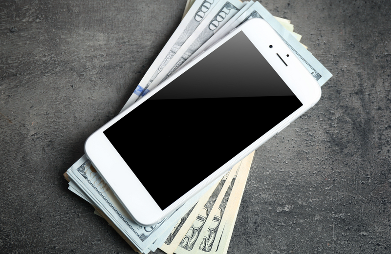 A white iPhone on top of a pile of American $100 notes on top of a slate surface. 