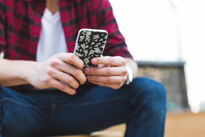 A man in a red plaid flannel, jeans and white shirt, holding a phone in both hands. The phone has a case with a paper aeroplane pattern.