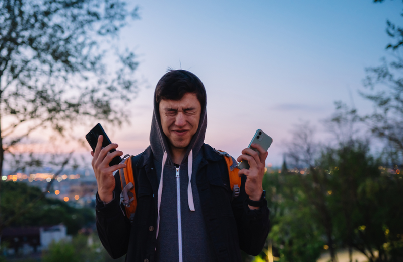 A man standing with two phones in his hand with a city skyline in the background at dusk. The man's face is scrunched.