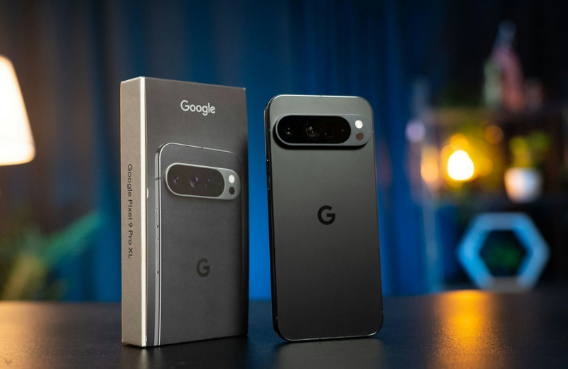 A black Google Pixel 9phone standing upright next to its manufacturers box on a black desk with lights out of focus in the background. 