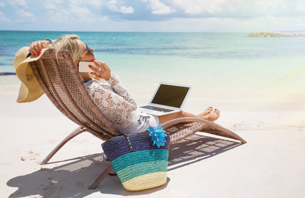 A blond woman talking on her phone as she lazes on the beach with a laptop resting on her lap.