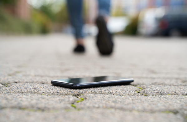 A black phone face down on a grey brick footpath. There is a person feet walking away out of focus in the background. They are wearing black shoes and blue jeans.