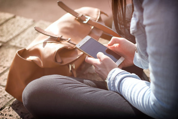 A girl crossed legged holding a phone with both hands and her tan handbag in front of her.