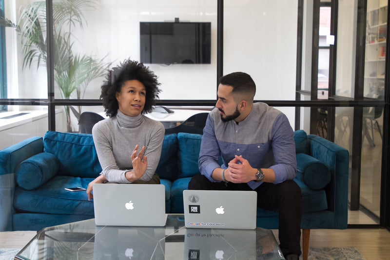 Two people sitting on a blue couch discussing business with MacBooks in front of them