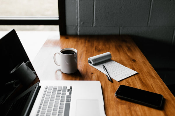 A Macbook on a wooden desk next to a notepad, iPhone and coffee mug.