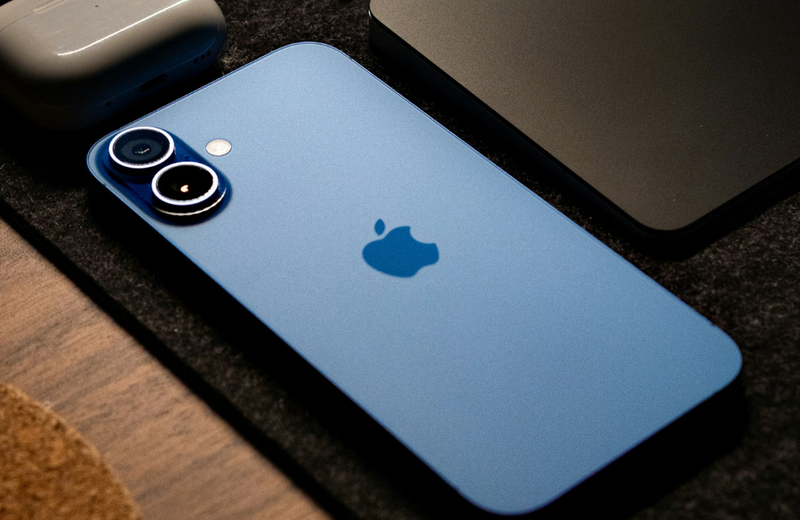 A blue iPhone 16 facedown on a desk with Air Pods above it being cut out of the frame.