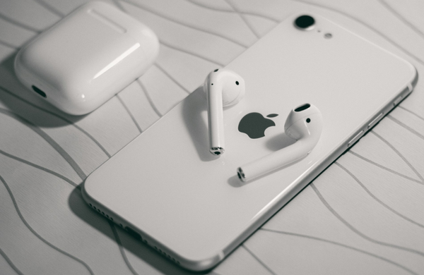 A white iPhone SE facedown on a white bench with unparallel lines. There are white Air Pod headphones on top of the phone and the casenext to it.  