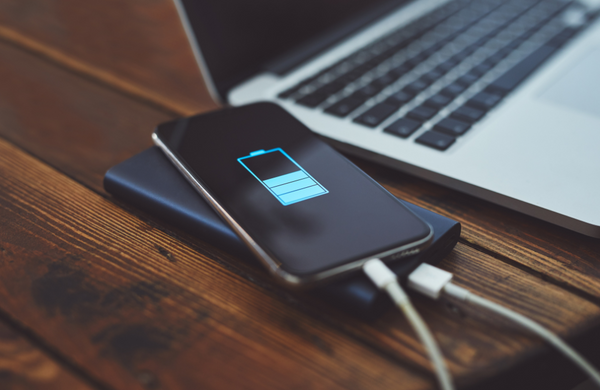 A phone plugged into a charging bank on a wooden table next to a laptop.